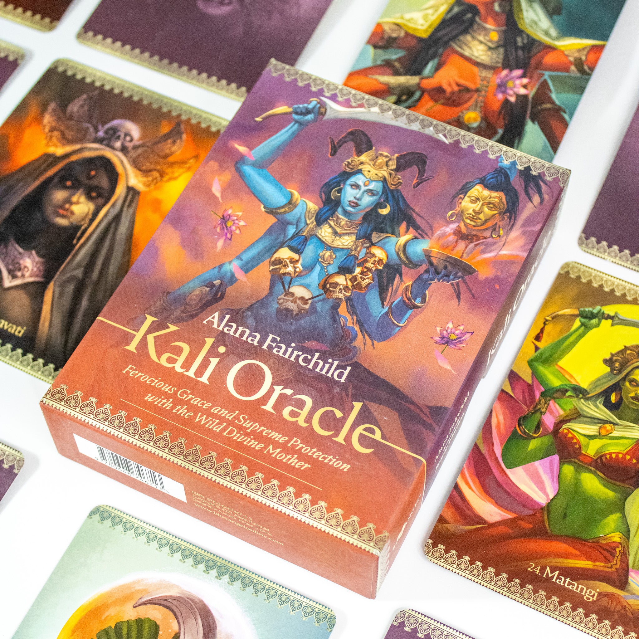Box of the Kali Oracle Deck by Alana Fairchild. Box is red and purple and shows a blue Kali with multiple arms wearing a necklace of skulls. She holds a blad. There is a severed head to her right. Other cards from the deck surround her.