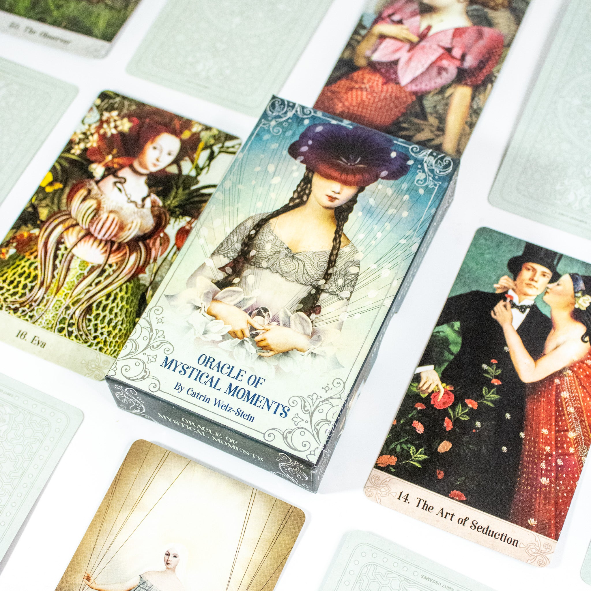 Box of Oracle of Mystical Moments Deck shows a woman with two brown braids. Her eyes are covered by a giant maroon flower. Other cards from the deck surround the box.