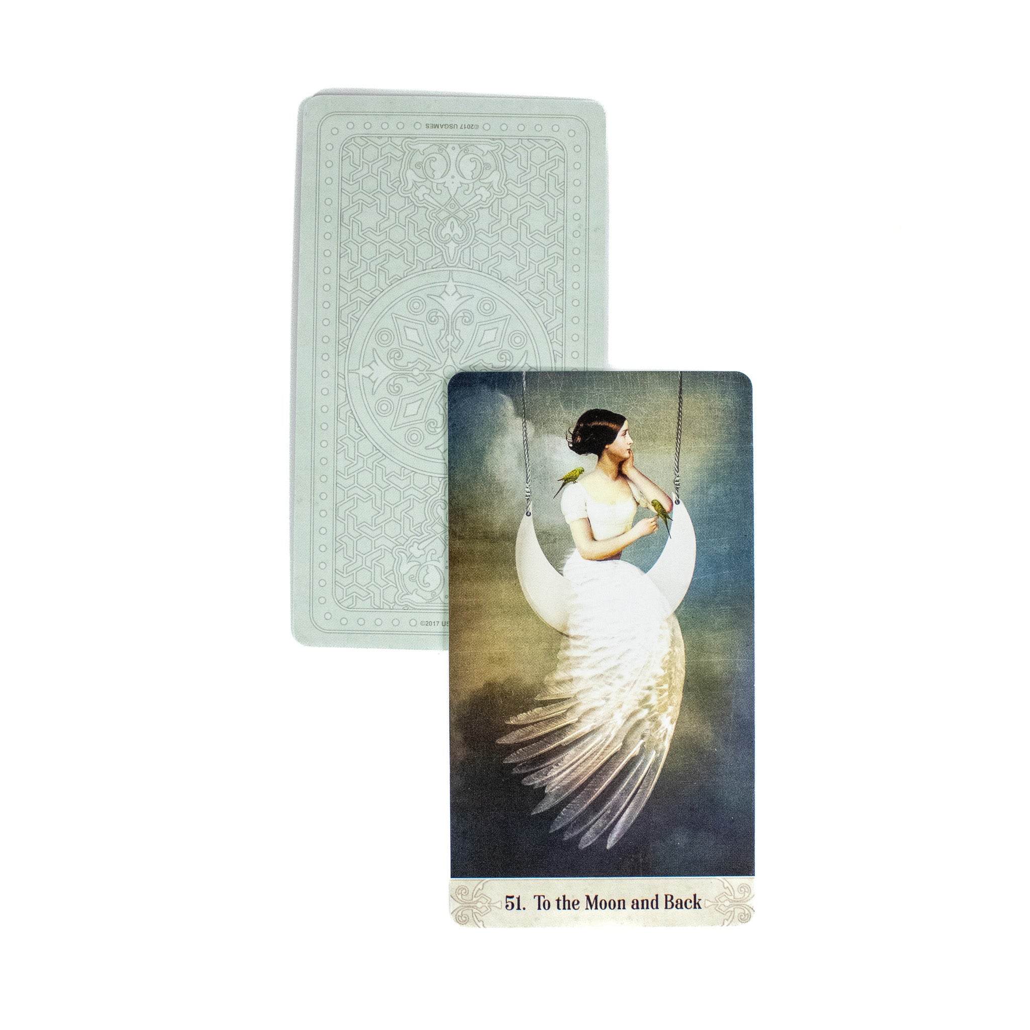 The To the Moon and Back oracle card shows a woman in a white dress sitting on a swing in the shape of a crescent moon. Her skirt is  made of feathers. A second card behind it shows the back of the deck, which is light green.