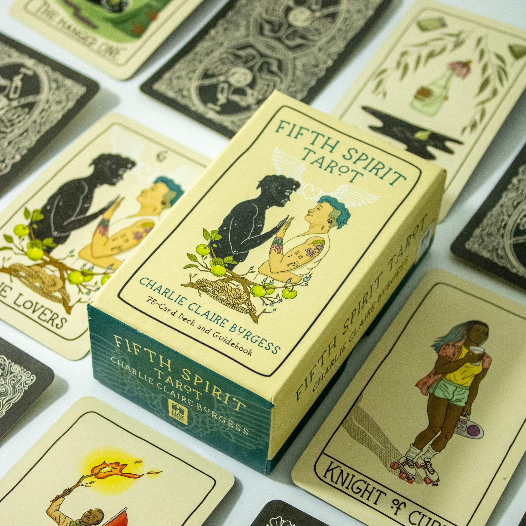 Fifth Spirit Tarot deck box shows two people palm to palm. One is in full color with short blue hair and tattoos, the other is a black outline with stars inside. There are apple twigs with apples below them and rope. Other cards from the deck surround the box.